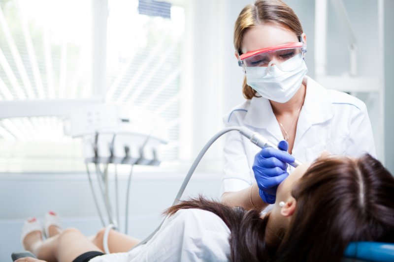 Dentist wearing eye shield and mask using tool on woman laying in dental chair